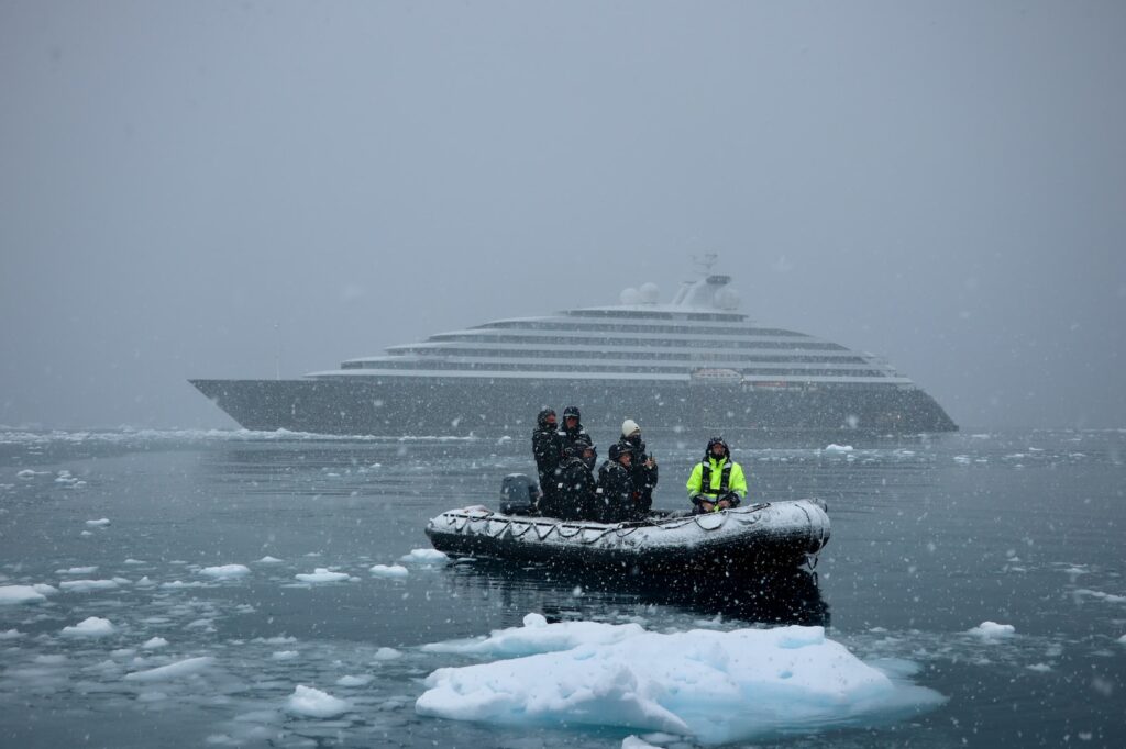 6 people in black jackets and one in yellow stand on a raft surrounded by ice with cruise ship in background 